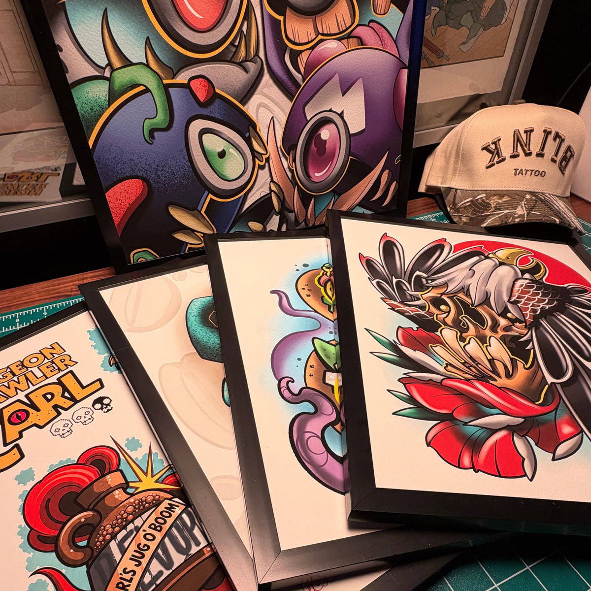 A collection of framed colorful cartoon and tattoo-style art prints arranged on a green cutting mat, with a cap and framed artwork in the background.